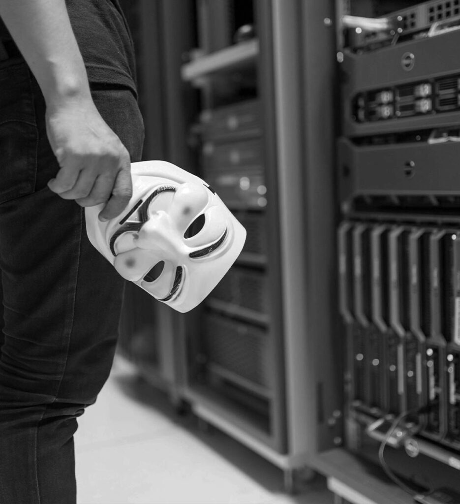 Hacker maszkot tartó kéz egy szerverteremben, ami a weboldalak feltörés elleni védelmét és a nyugodt, havidíjas WordPress karbantartást jelképezi.